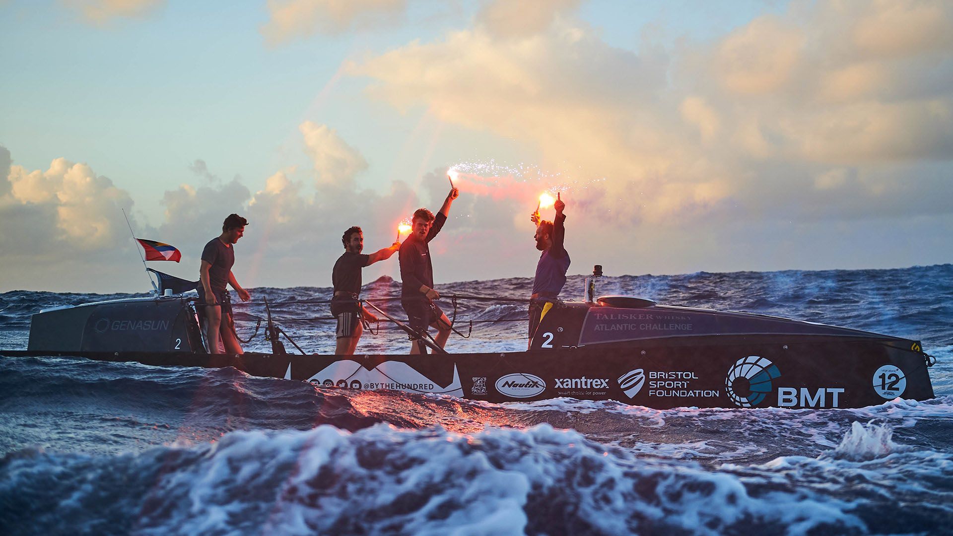 For men in wetsuits in rough ocean water, holding flares aloft and piloting a sporty boat.